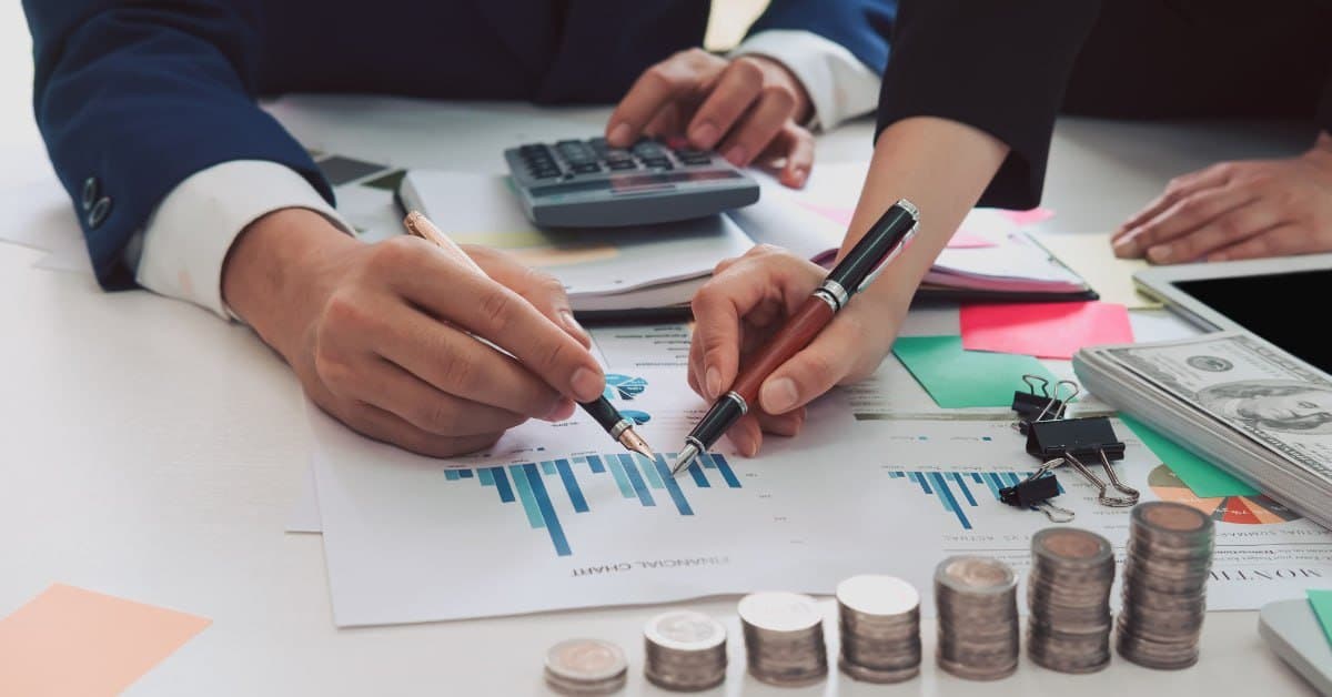 Two marketing professionals reviewing printed financial charts and performance data at a desk with a calculator and stacked coins