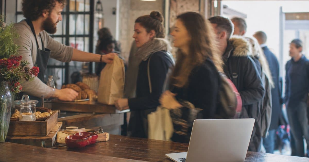 A busy shop owner serving a queue of customers while a laptop sits unattended on the counter